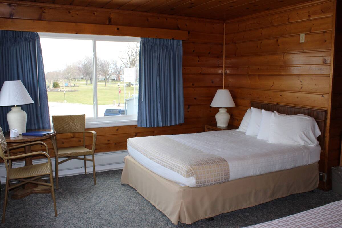 A wooden cabin room with a bed, two chairs, a table, and a window with curtains.