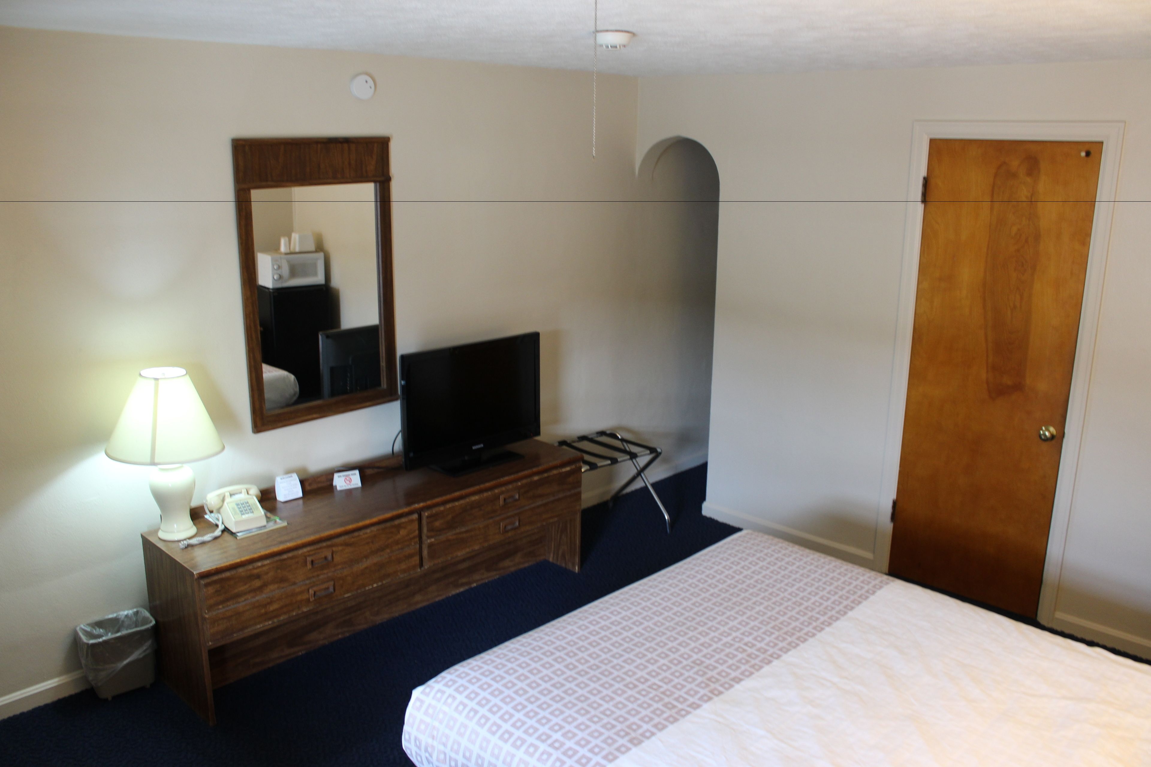 Habitación de hotel con cama, cómoda con espejo, televisor, lámpara, teléfono y puerta.