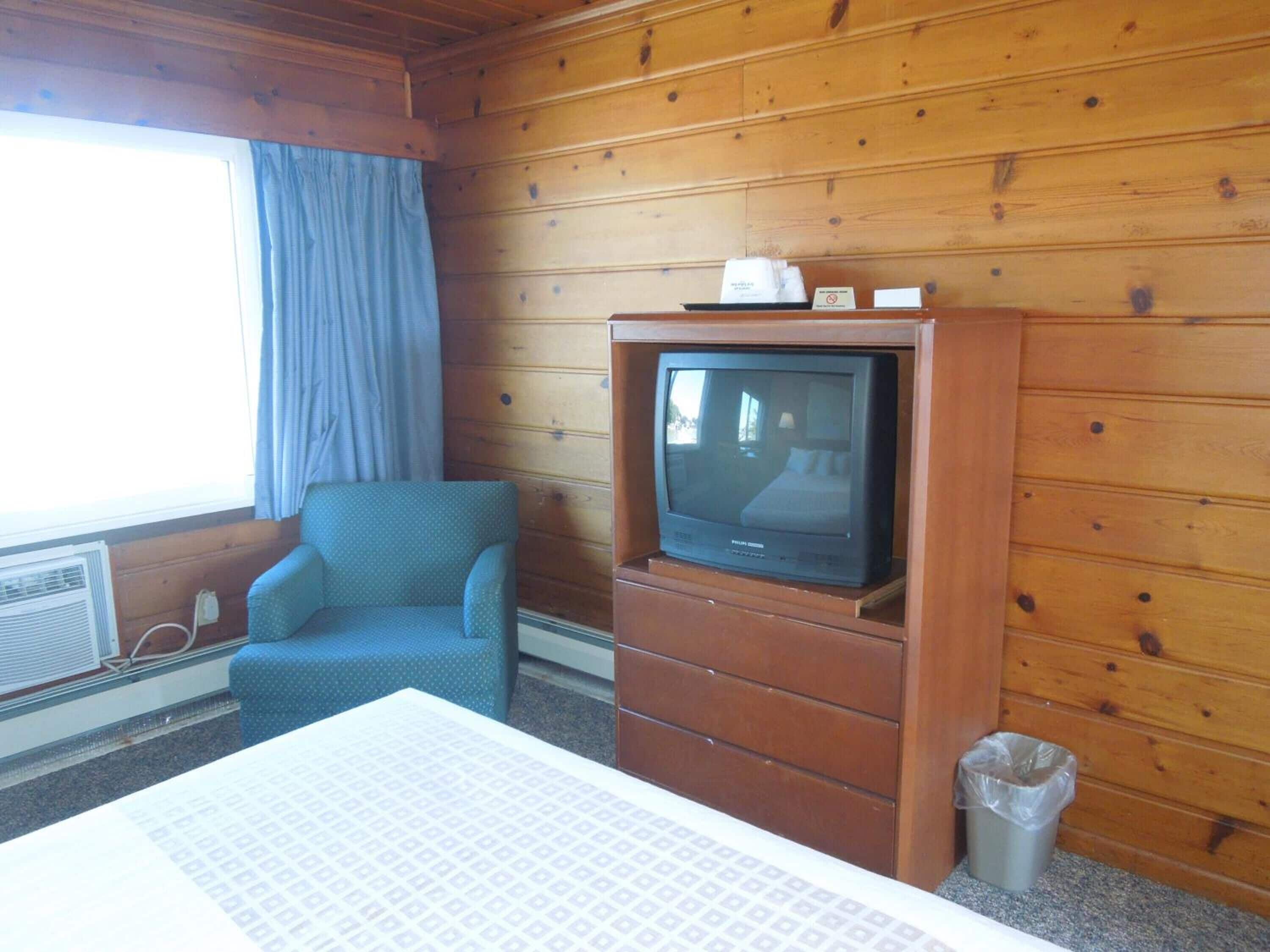 A hotel room with a wooden paneled wall, a single bed, a blue armchair, a television on a wooden cabinet, and a window with curtains.