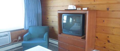 A hotel room with a wooden paneled wall, a single bed, a blue armchair, a television on a wooden cabinet, and a window with curtains.