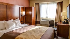 A hotel room with a large bed, a desk, a chair, a lamp, an air conditioning unit, and a window with curtains. - Ramada by Wyndham New York Times Square West (New York)