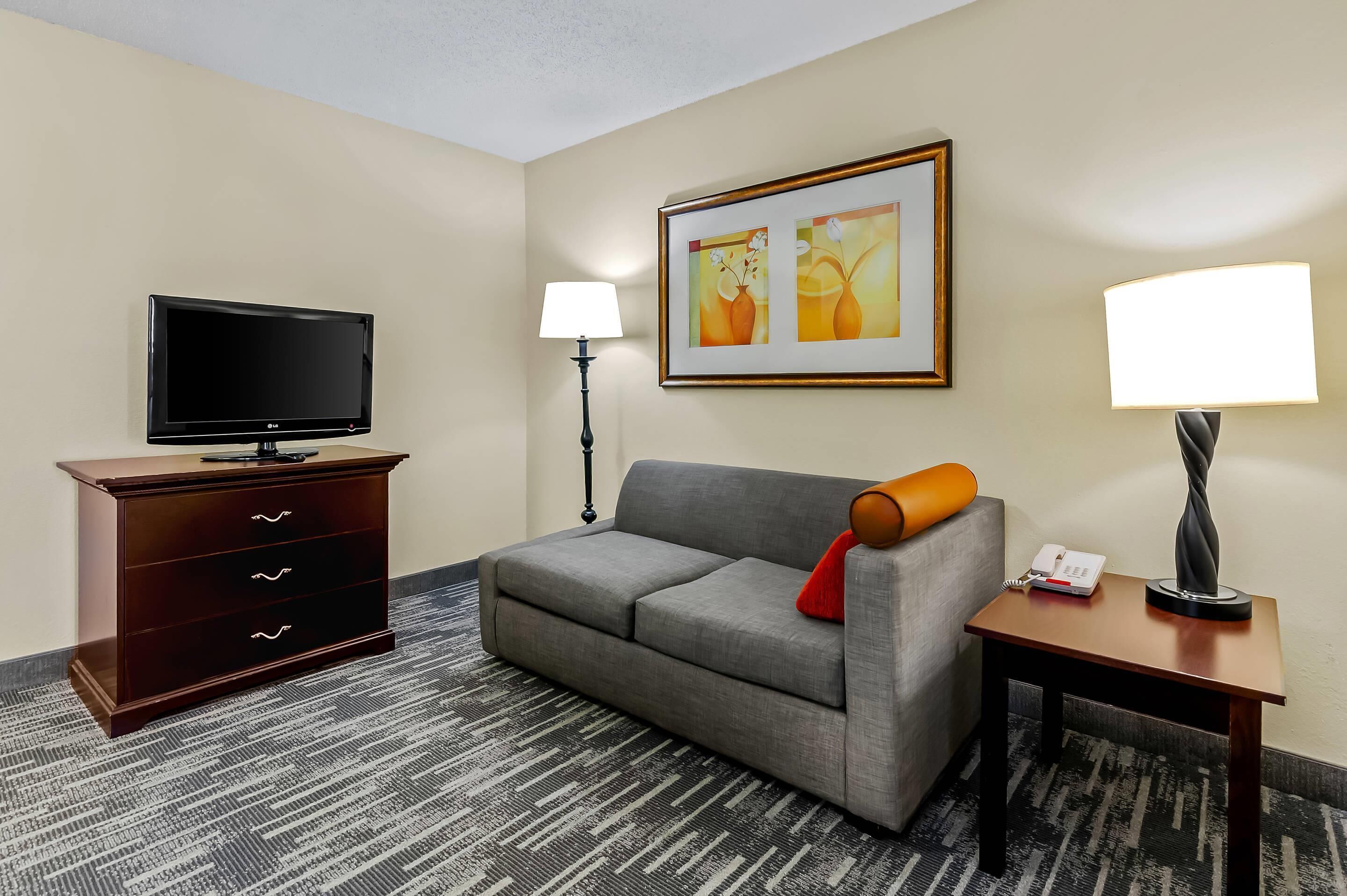 A hotel room with a flat-screen TV, a wooden dresser, a gray sofa with a red pillow, two wall-mounted lamps, and a framed artwork above the sofa.