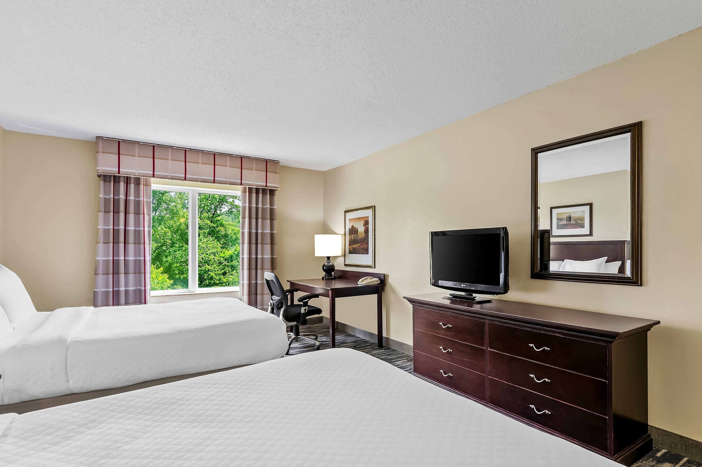 Une chambre d’hôtel comprenant un lit, un bureau avec une chaise, une télévision, une commode et une fenêtre avec des rideaux.