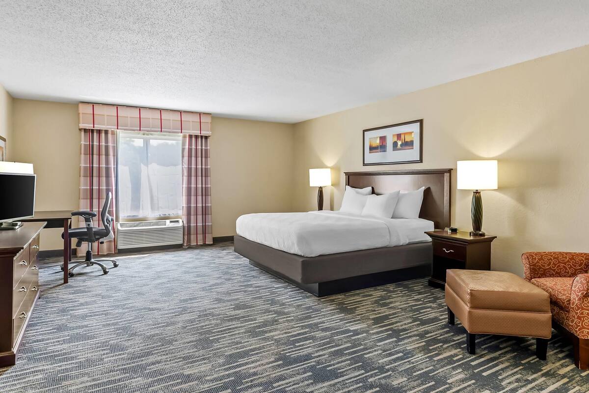A hotel room with a bed, two bedside lamps, a desk with a chair, a TV, and a window with curtains.
