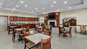 A spacious dining area with wooden tables and chairs, a kitchen in the background, and a flat-screen TV mounted above the fireplace.