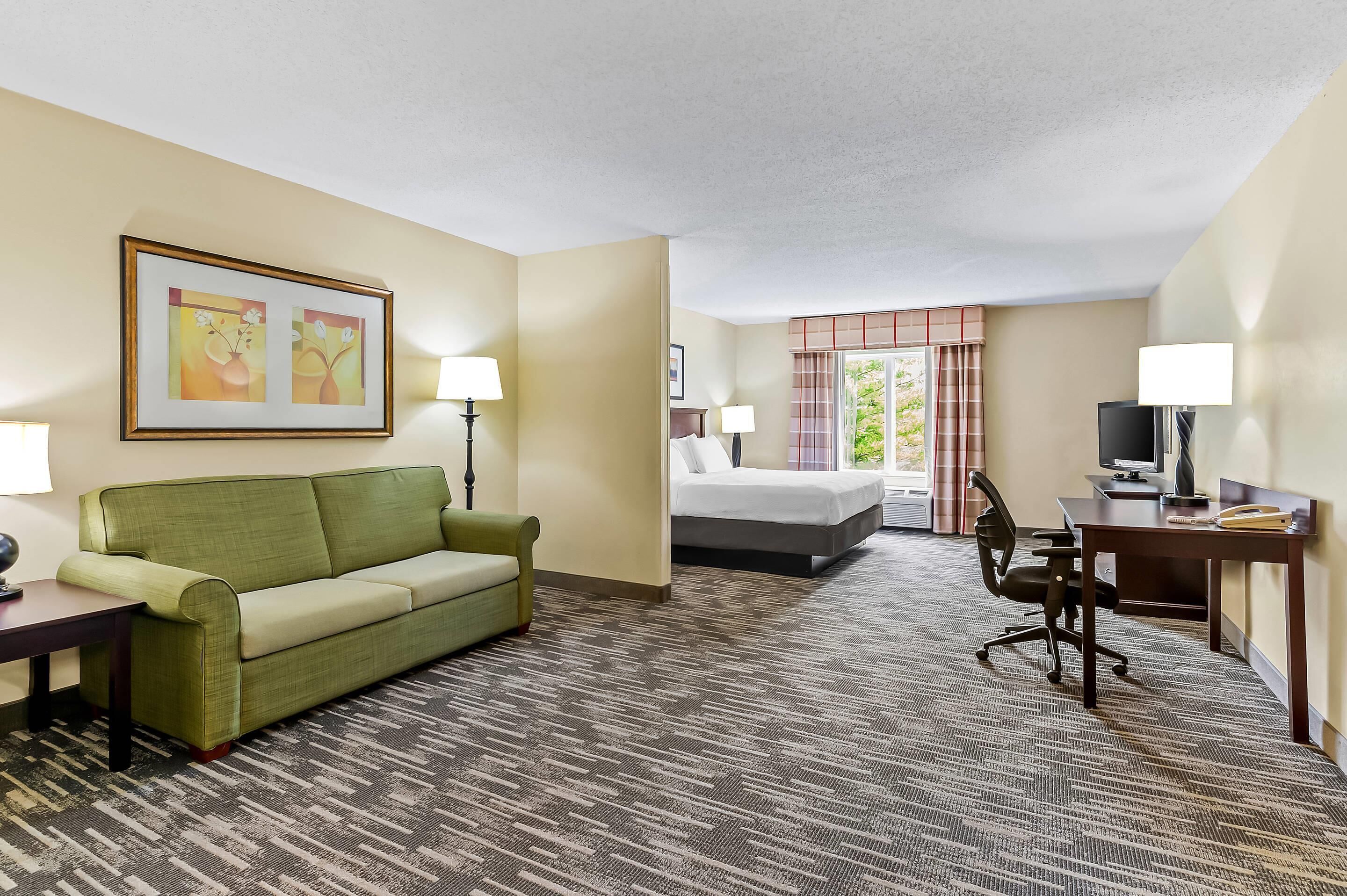 A hotel room with a green sofa, a bed, a desk with a computer, and a TV. There are two lamps on the desk and a window with curtains.
