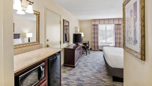 Habitación de hotel con una cama grande, un escritorio con televisor, cómoda, espejo y ventana con cortinas.