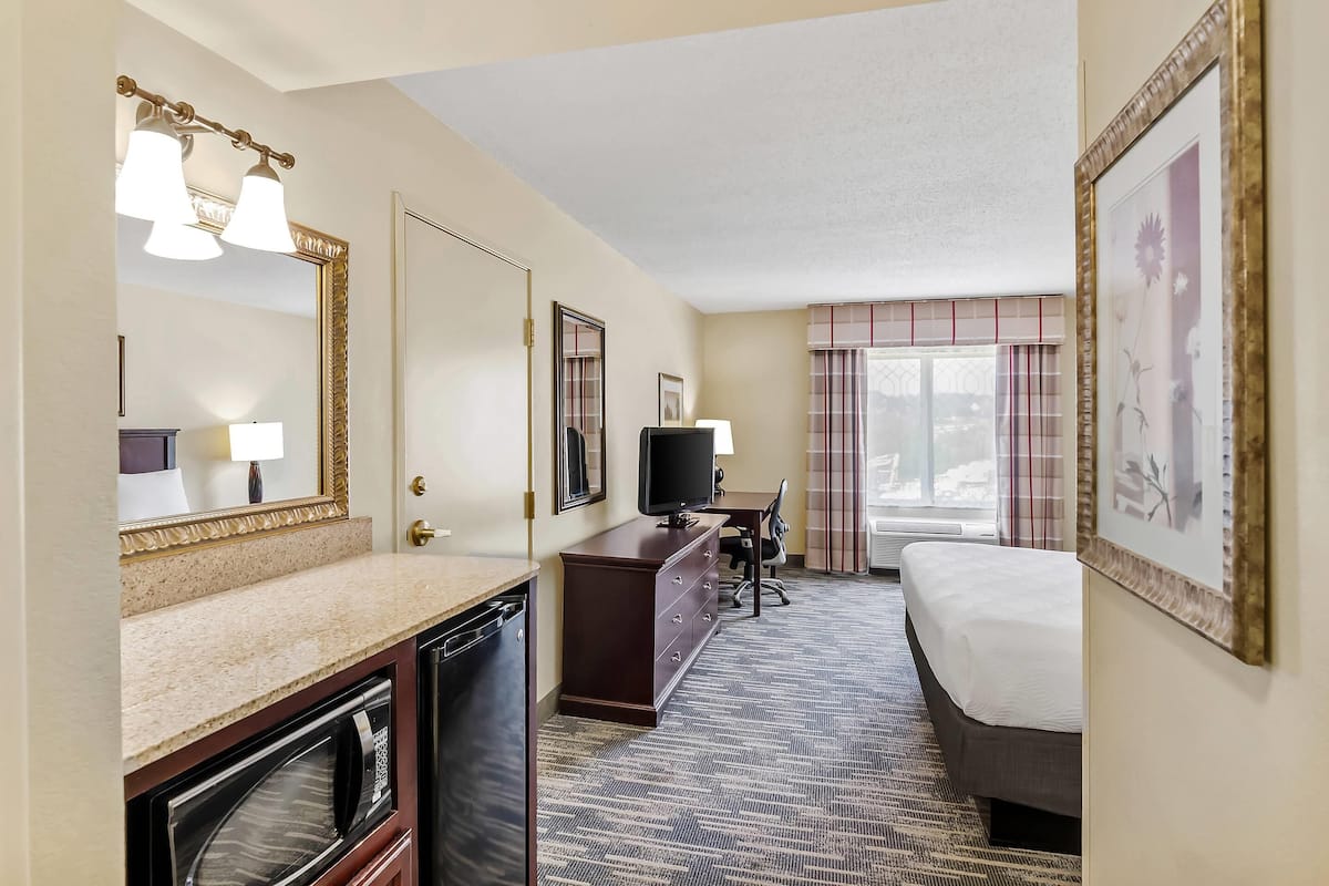 A hotel room with a large bed, a desk with a television, a dresser, a mirror, and a window with curtains.