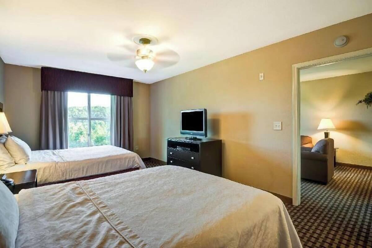 Habitación de hotel con dos camas, un televisor sobre un tocador, un ventilador de techo y vistas al exterior a través de la ventana.