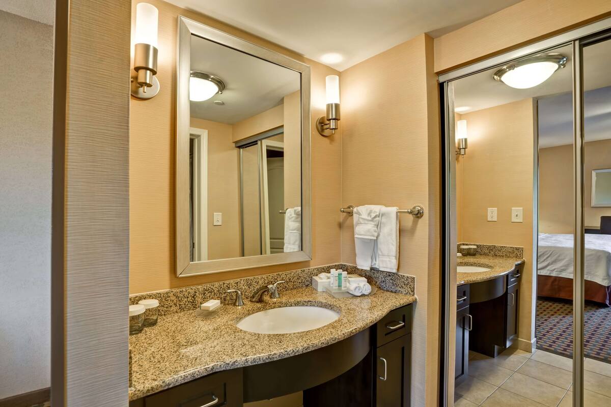 Un baño de hotel con lavamanos de encimera de granito, un espejo grande y un tocador independiente con lavamanos.