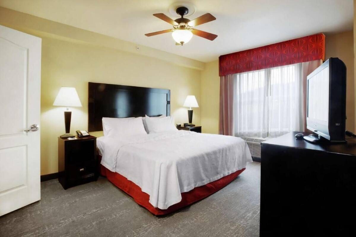 Habitación de hotel con cama, dos mesitas de noche con lámparas, ventilador de techo, televisor y ventana con cortinas.