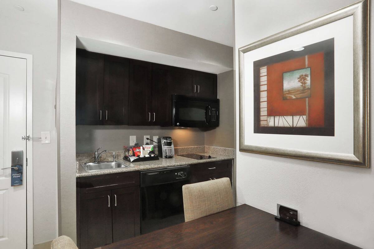 Una cocina compacta con armarios oscuros, un microondas, una cafetera y un lavamanos. Hay un cuadro en la pared y una mesa de comedor con una silla.