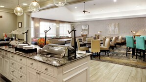 Une salle à manger d’hôtel avec un buffet, plusieurs tables, des chaises et un ventilateur de plafond.