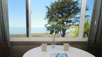 Una mesa redonda con dos tazas blancas y una revista, ubicada cerca de una ventana que ofrece vista al océano y un árbol.