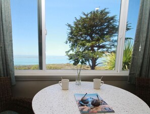 A round table with two white mugs and a magazine, positioned by a window with a view of the ocean and a tree.