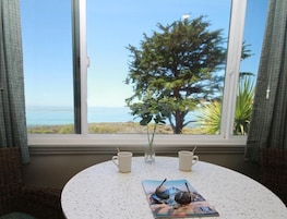 Una mesa redonda con dos tazas blancas y una revista, ubicada cerca de una ventana que ofrece vista al océano y un árbol.