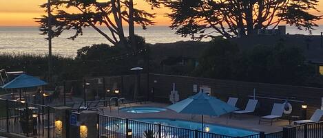 A pool area with lounge chairs and umbrellas, surrounded by a fence, overlooking a body of water at sunset.