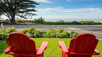 Two red Adirondack chairs facing a view of the ocean with a grassy area and a tree in the foreground.