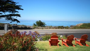 A coastal view with three red Adirondack chairs facing the ocean, a stone pillar, and a variety of shrubs and flowers. - Castle Inn (Cambria)