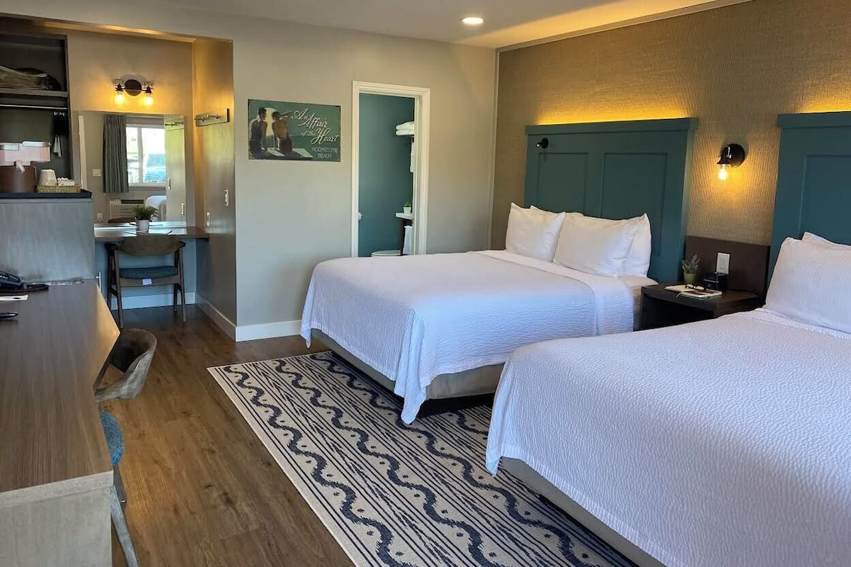 A hotel room with two beds, a desk, a chair, a patterned rug, and a view of the beach.