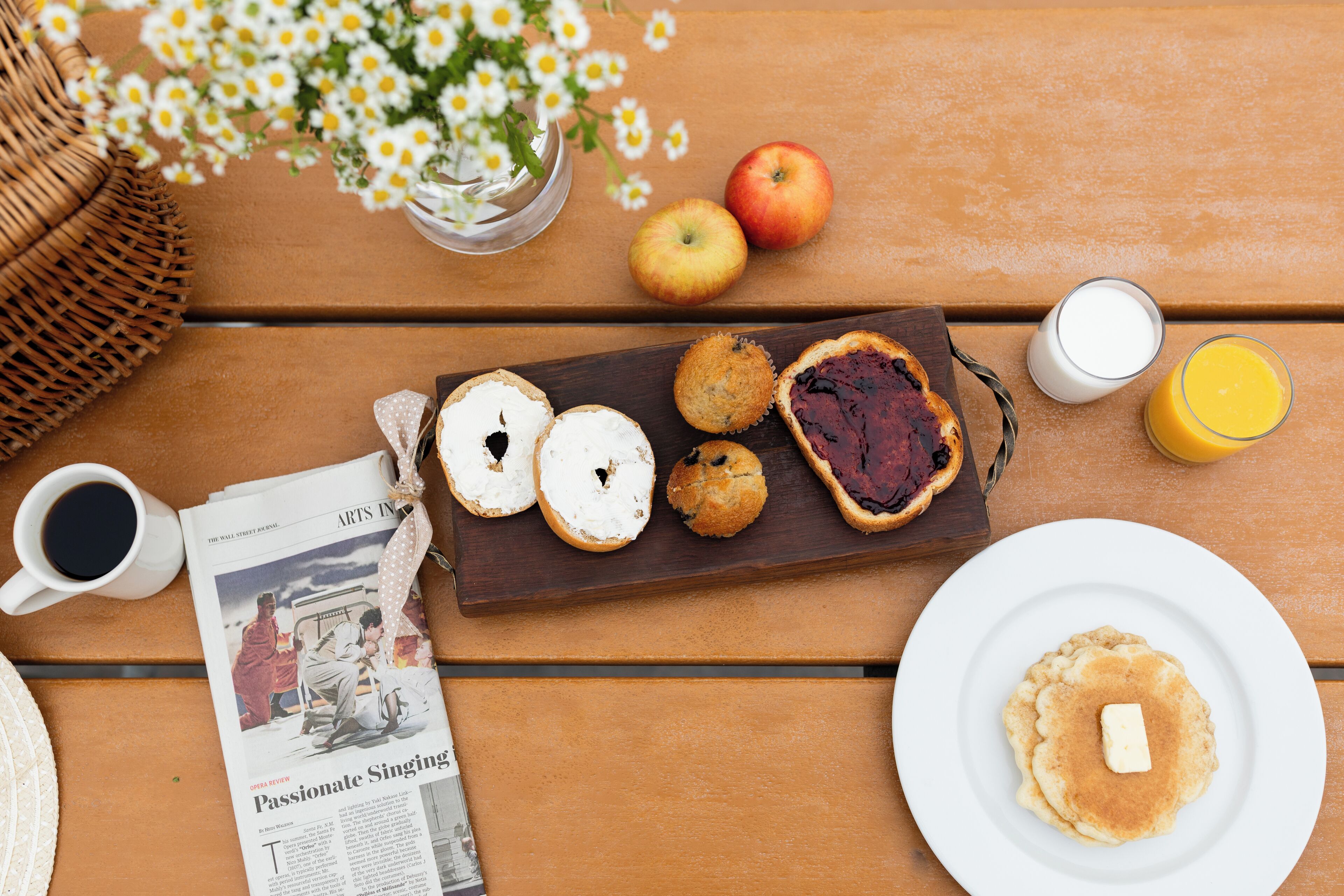Ein Holztisch mit Frühstücksangeboten, darunter Gebäck, eine Zeitung und eine Vase mit Blumen.