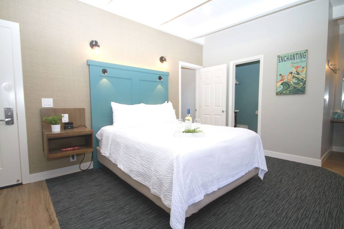 A hotel room with a bed, a bedside table with a plant, a wall-mounted light, and a door.