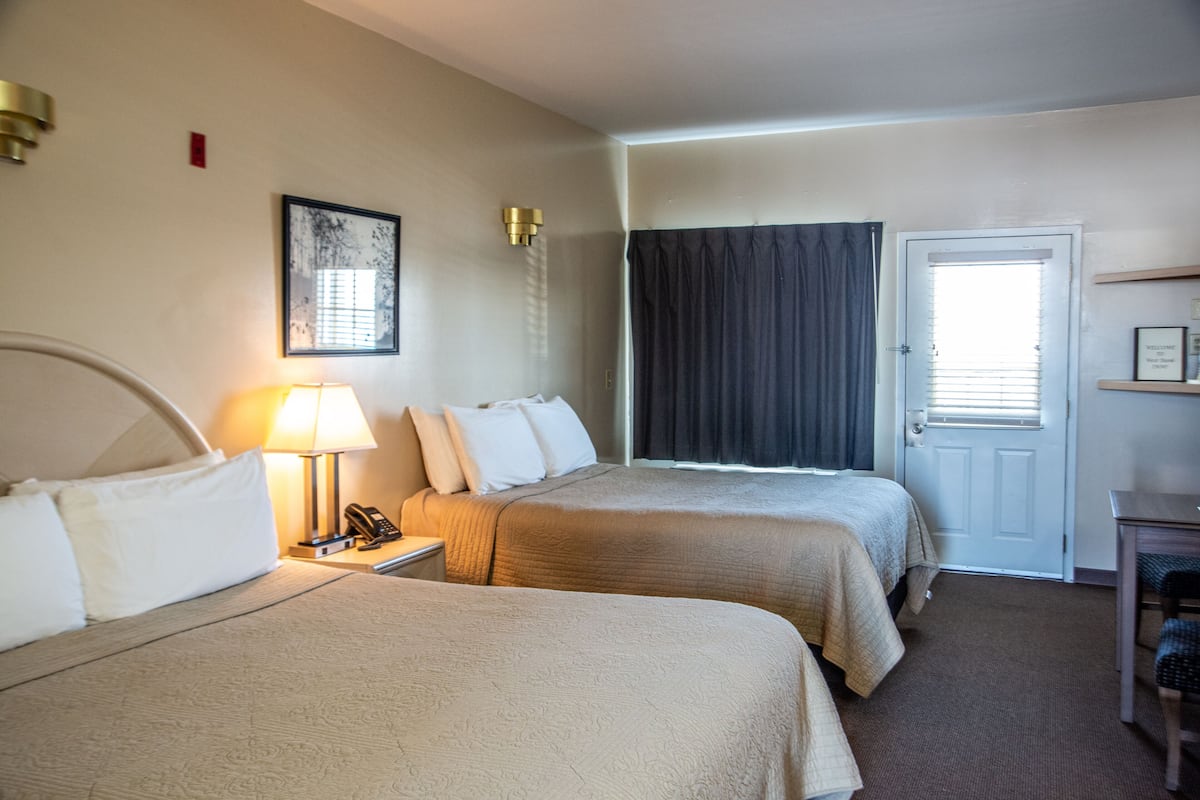 A hotel room with two beds, a bedside table with a lamp, a telephone, a door with a window, and a small table with a chair.