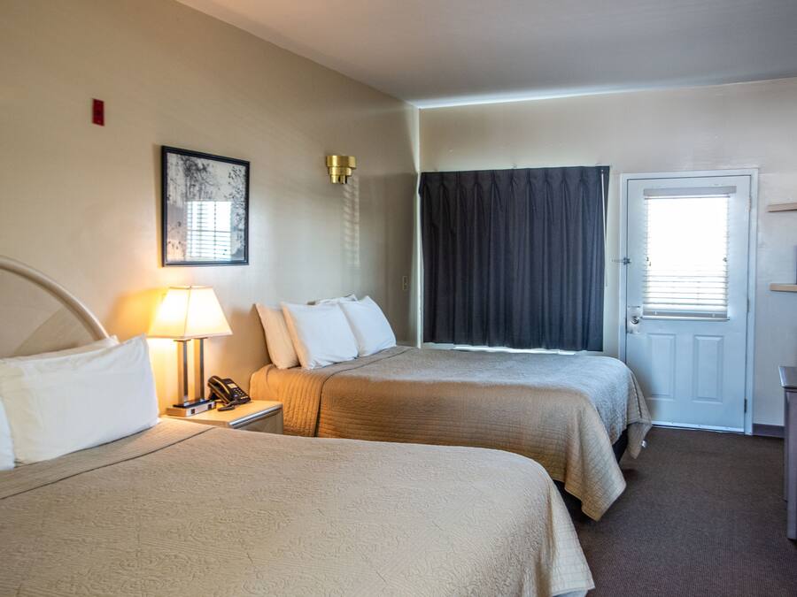 A hotel room with two beds, a bedside table with a lamp, a telephone, a door with a window, and a small table with a chair.