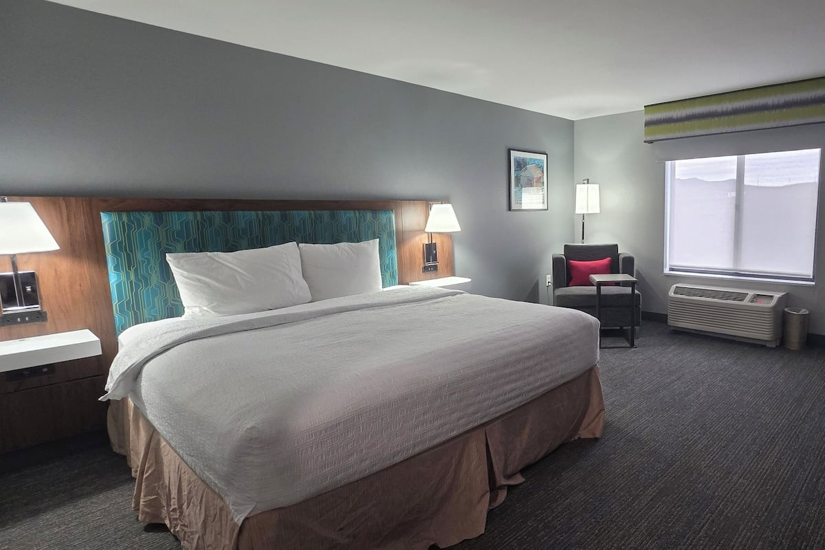 A hotel room with a large bed, two bedside tables with lamps, a chair, a desk with a computer, and a window with blinds.