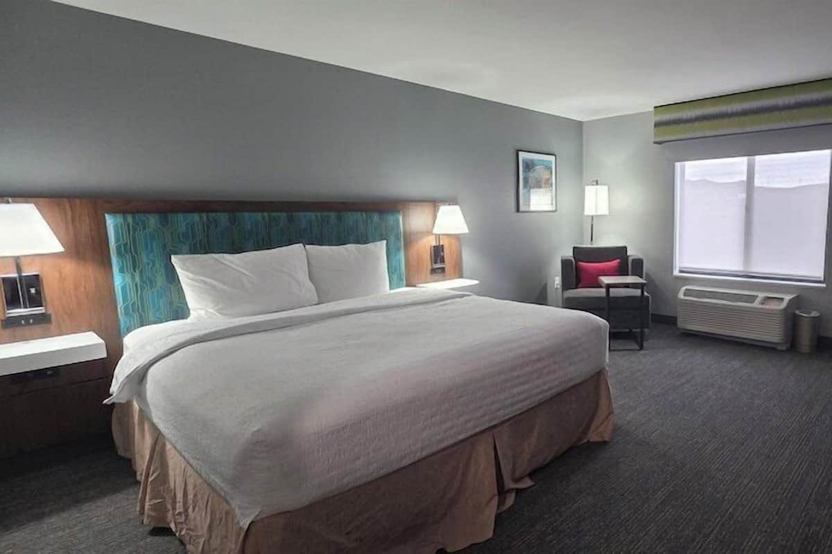 A hotel room with a large bed, bedside lamps, a desk, a chair, a window with blinds, and a radiator.
