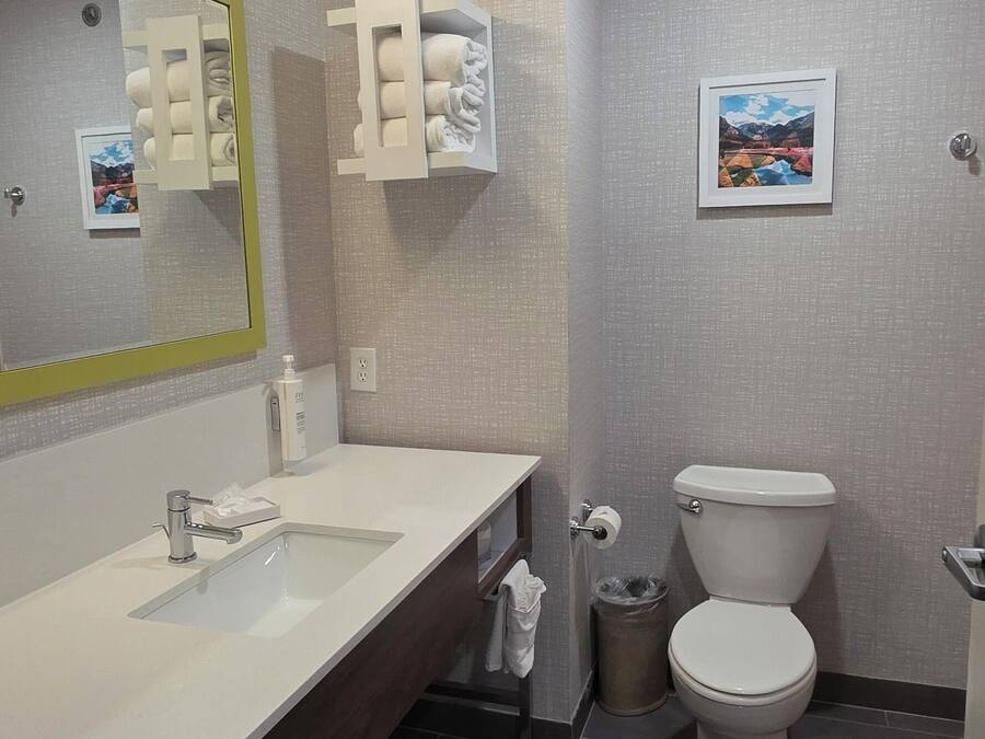 A modern bathroom with a white sink, mirror, and toilet. There are towel racks and a trash can.