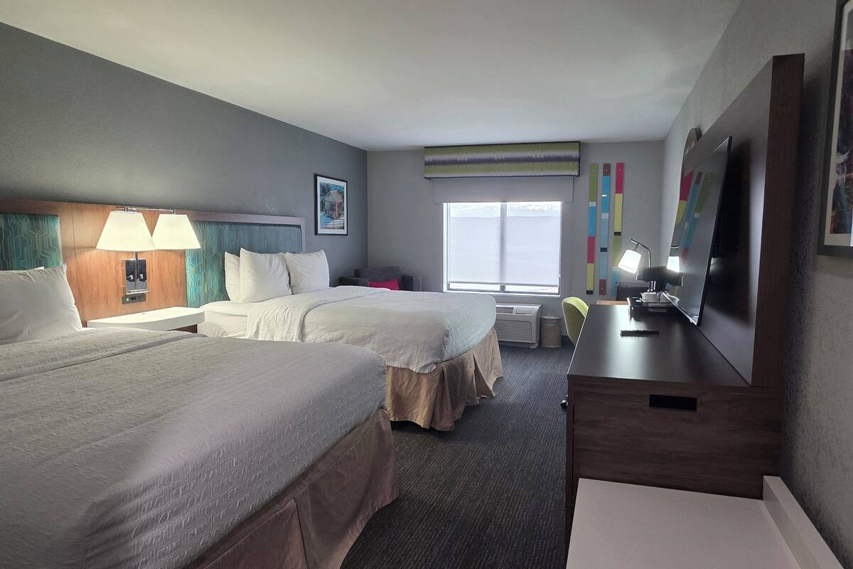 A hotel room with two beds, a desk, a TV, and a window with blinds.