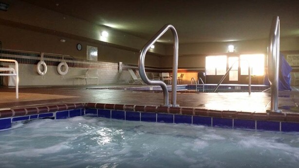 Una piscina interior con una escalera metálica, sillas de descanso y suelo de baldosas.