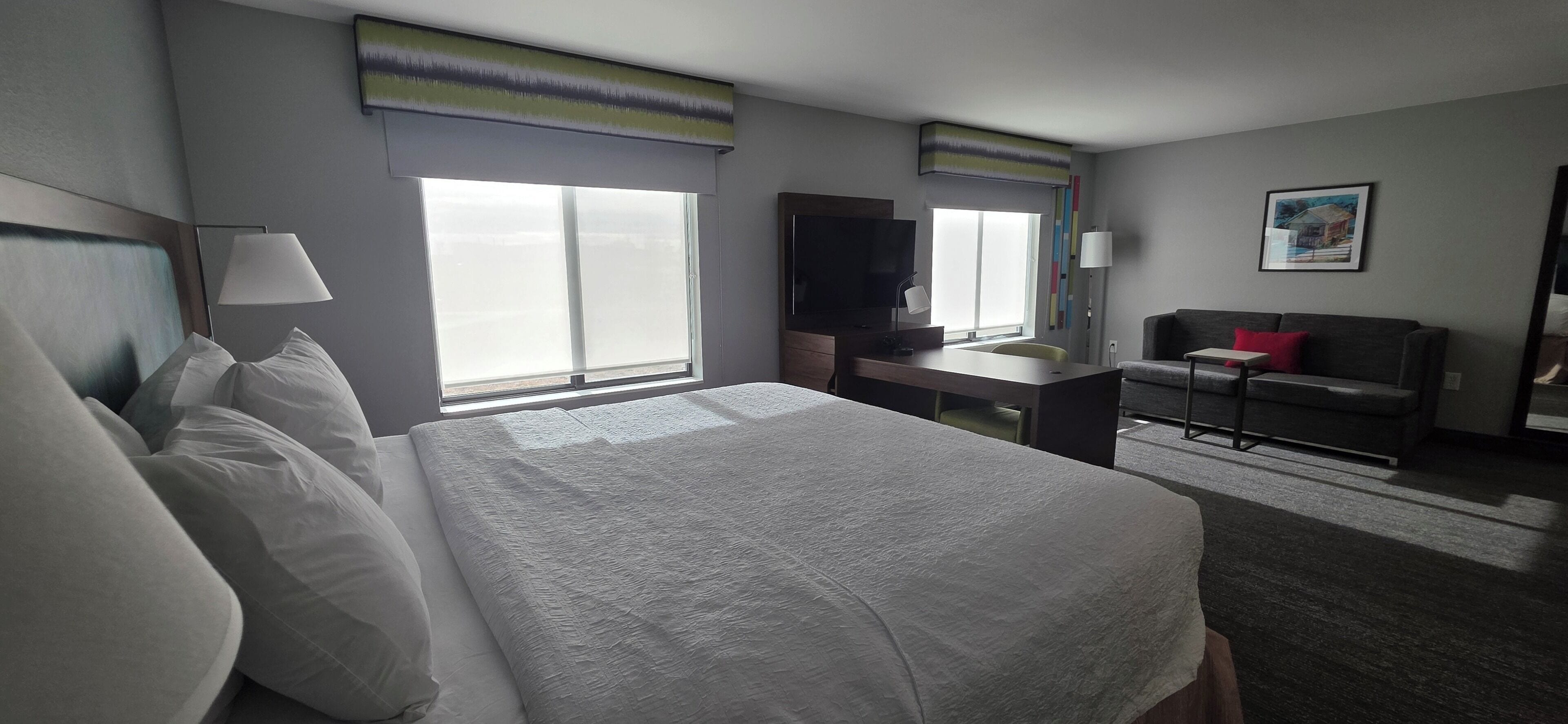 A hotel room with a large bed, a desk with a flat-screen TV, a chair, a small table, and a window with blinds.