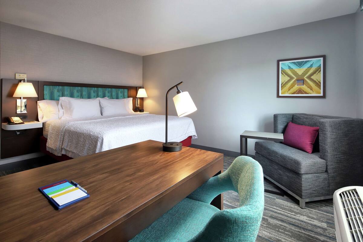A hotel room with a bed, bedside tables with lamps, a desk with a tablet, a sofa, and a wall-mounted picture.