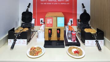 Una estació de gofres amb dues planxes per a gofres, un cartell que diu 'CRISP & DELIGHTFUL HAPPY WAFFLE, HAPPY DAY YOU ARE GOING TO LOVE IT A WAFFLE LOT!', i dues plates de gofres amb ingredients.