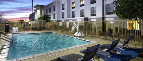 A swimming pool with lounge chairs and a fenced area, surrounded by a building with multiple windows.