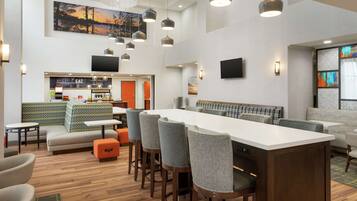 Un hall d’hôtel moderne doté d’une longue table commune, d’un bar, de coins salon et d’un écran de télévision.