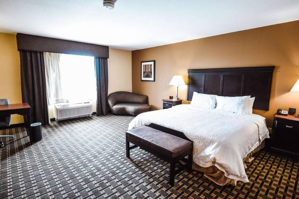 Quarto de hotel com uma cama grande, uma escrivaninha, uma cadeira, um banco, uma mesa de cabeceira, um abajur e um quadro na parede.