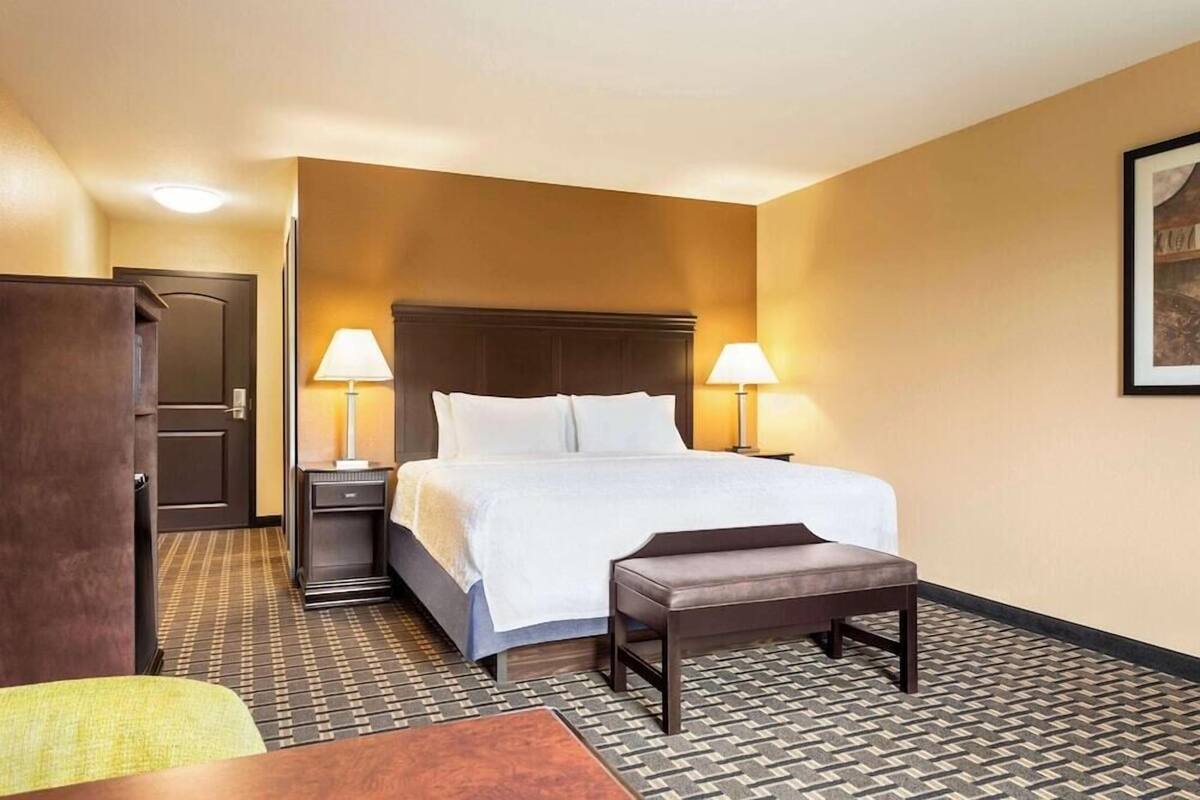 A hotel room with a bed, bedside lamps, a bench, a desk, and a door.