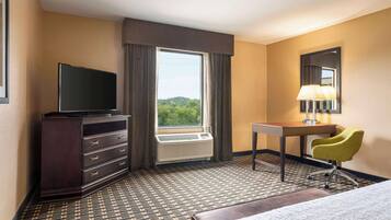 A hotel room with a large window, a flat-screen TV, a desk with a lamp, a green chair, and a bed.