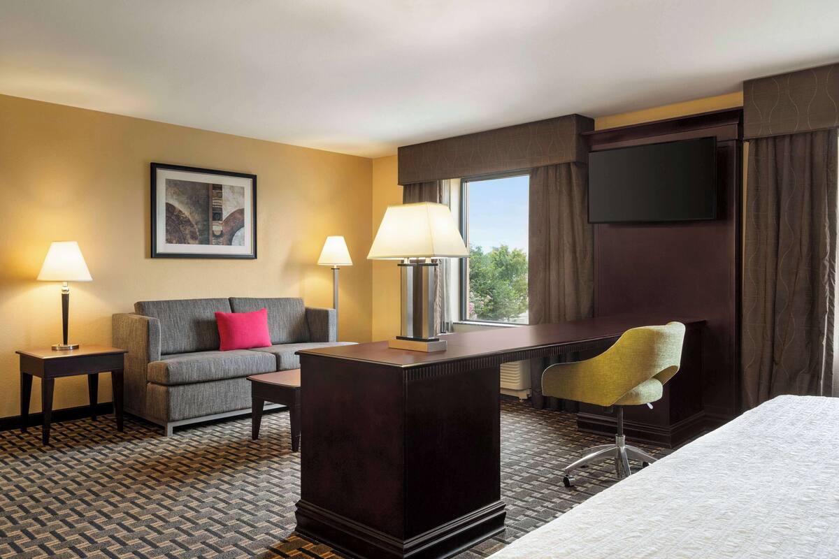 Quarto de hotel com cama, escrivaninha, sofá, duas luminárias, televisão de tela plana e janela com cortinas.
