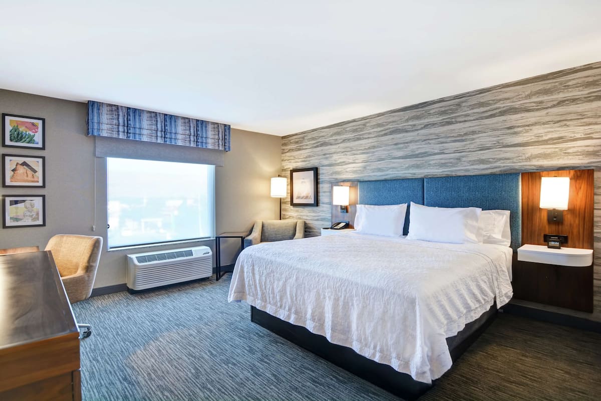 A hotel room with a large bed, a desk, a chair, a window with a view, and a wall-mounted air conditioner.