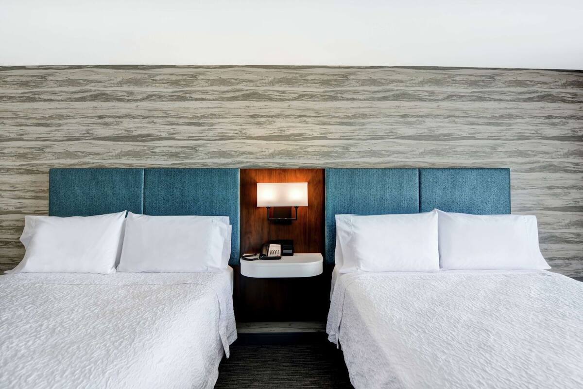 A hotel room with two beds, a nightstand, a lamp, and a small sink.