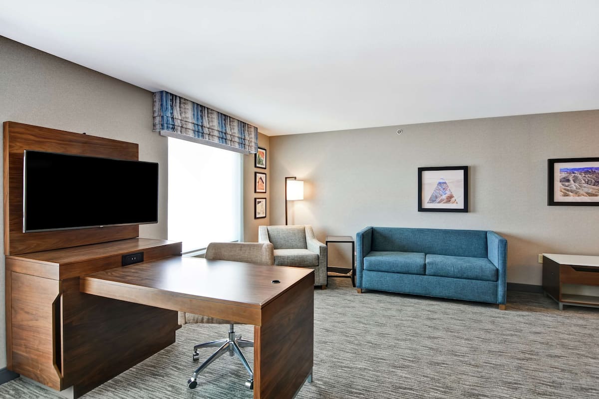 A hotel room with a wooden desk, a flat-screen TV, a blue sofa, a small table, and framed pictures on the wall.