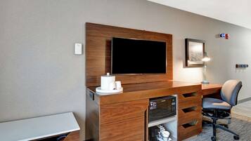 A hotel room with a wooden desk, a flat-screen TV, a lamp, a chair, and a small shelf with a coffee maker and cups.