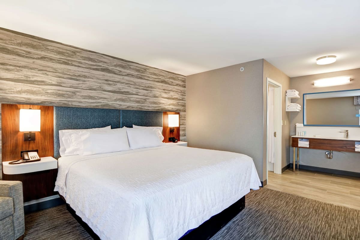 A hotel room with a large bed, bedside tables with lamps, a wooden headboard, a bathroom with a mirror, and a wooden floor.