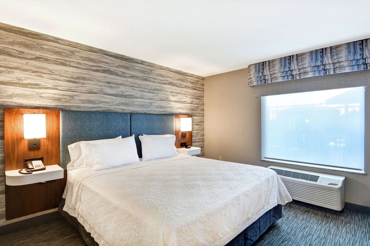 A hotel room with a large bed, two bedside tables with lamps, a built-in heater, and a large window with a view.