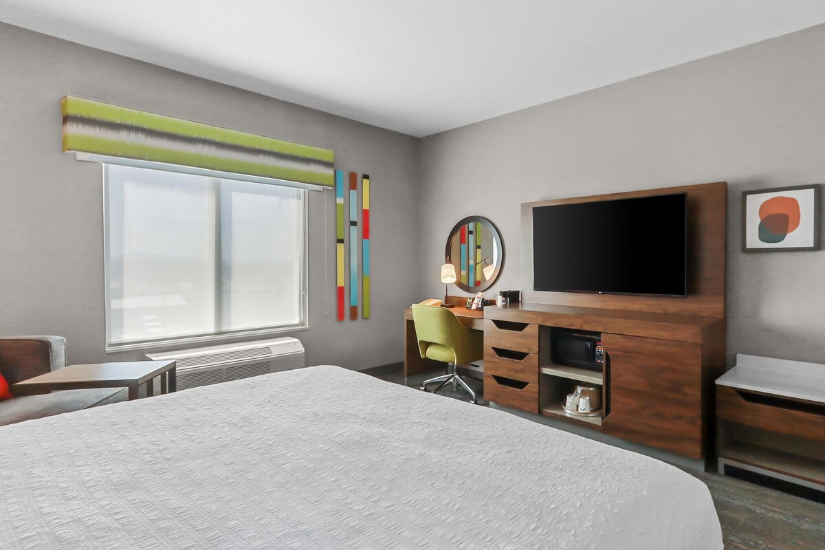 A hotel room with a large bed, a desk with a chair, a television, and a window with blinds.