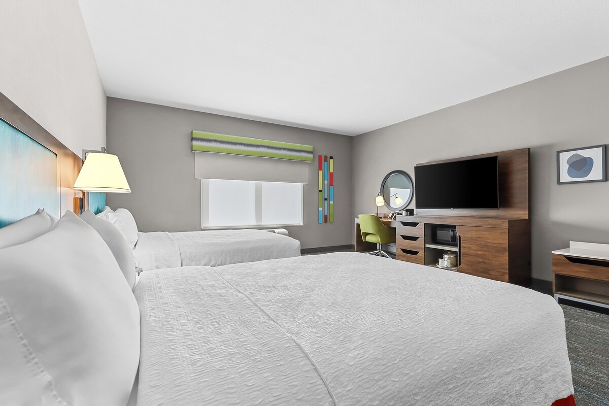 A hotel room with two beds, a TV, a desk, a lamp, and a mirror.